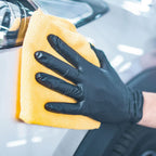 Eine hand mit schwarzem hygienstar safe light nitrilhandschuh wischt sanft eine autofläche mit gelbem mikrofasertuch