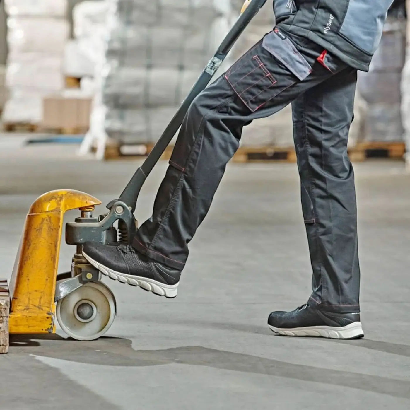 Schwarze arbeitshose mit mehreren taschen und roten nähte, getragen von einem arbeiter mit gelbem palettenhubwagen, kombiniert mit wica® lanzo 31540 halbschuhen s1p esd sra